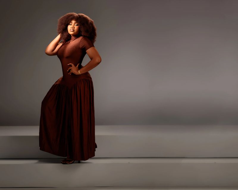 An African woman with voluminous afro hair poses elegantly in a studio. She wears a brown flowing dress, hand on her waist and the other touching her hair, radiating style, strength, and femininity.