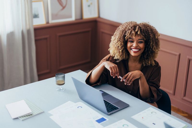 Professional workspace with a cheerful individual using a laptop, surrounded by documents and natural light, emanating a relaxed yet productive ambiance in a well-decorated setting.