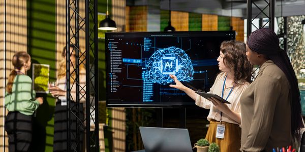 Two women discuss AI brain model on screen in modern office.