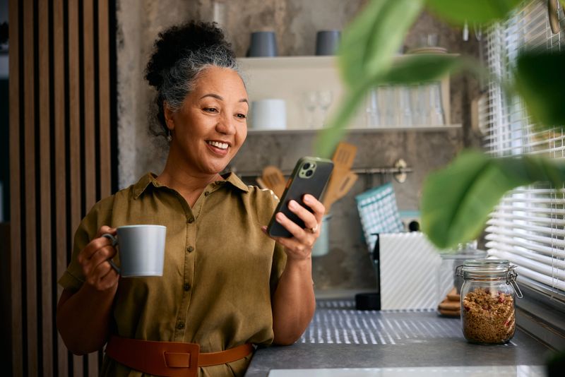 Happy mature woman holding a cup of coffee and using a mobile phone in the kitchen, smiling while looking at the screen, enjoying a relaxed morning moment at home