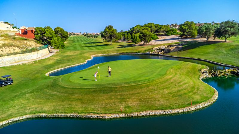 Two golfers stand on a lush green surrounded by water, enjoying a sunny day at a picturesque golf course. Trees and distant houses complete the serene setting
