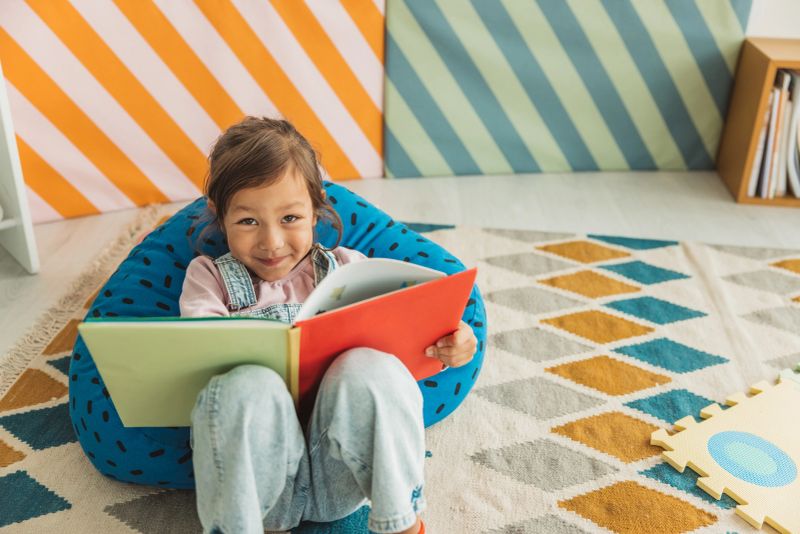 An adorable preschool age little girl smiles as she laying down in her preschool classroom and reading book.