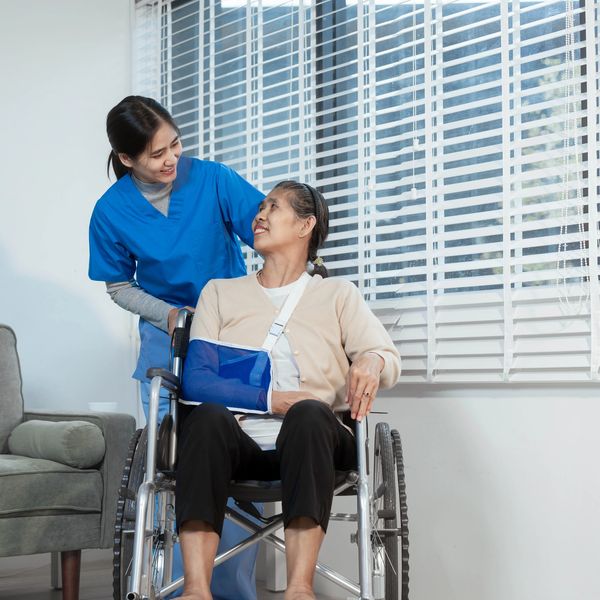 Caregiver in blue scrubs assisting a woman in a wheelchair with an arm injury.