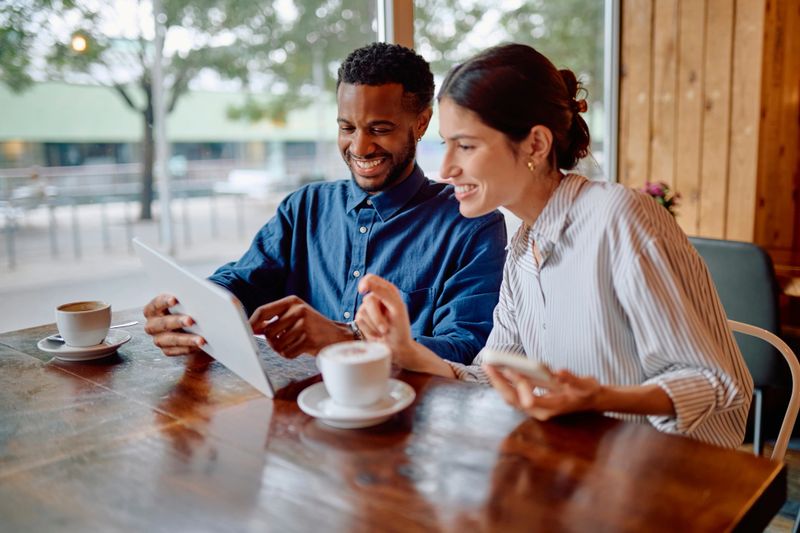 Two young colleagues smiling and enjoying a coffee break while using a digital tablet in a cozy cafe, sharing ideas and connecting