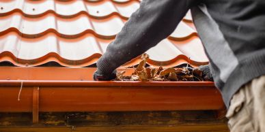 Person cleaning leaves from a red roof gutter.