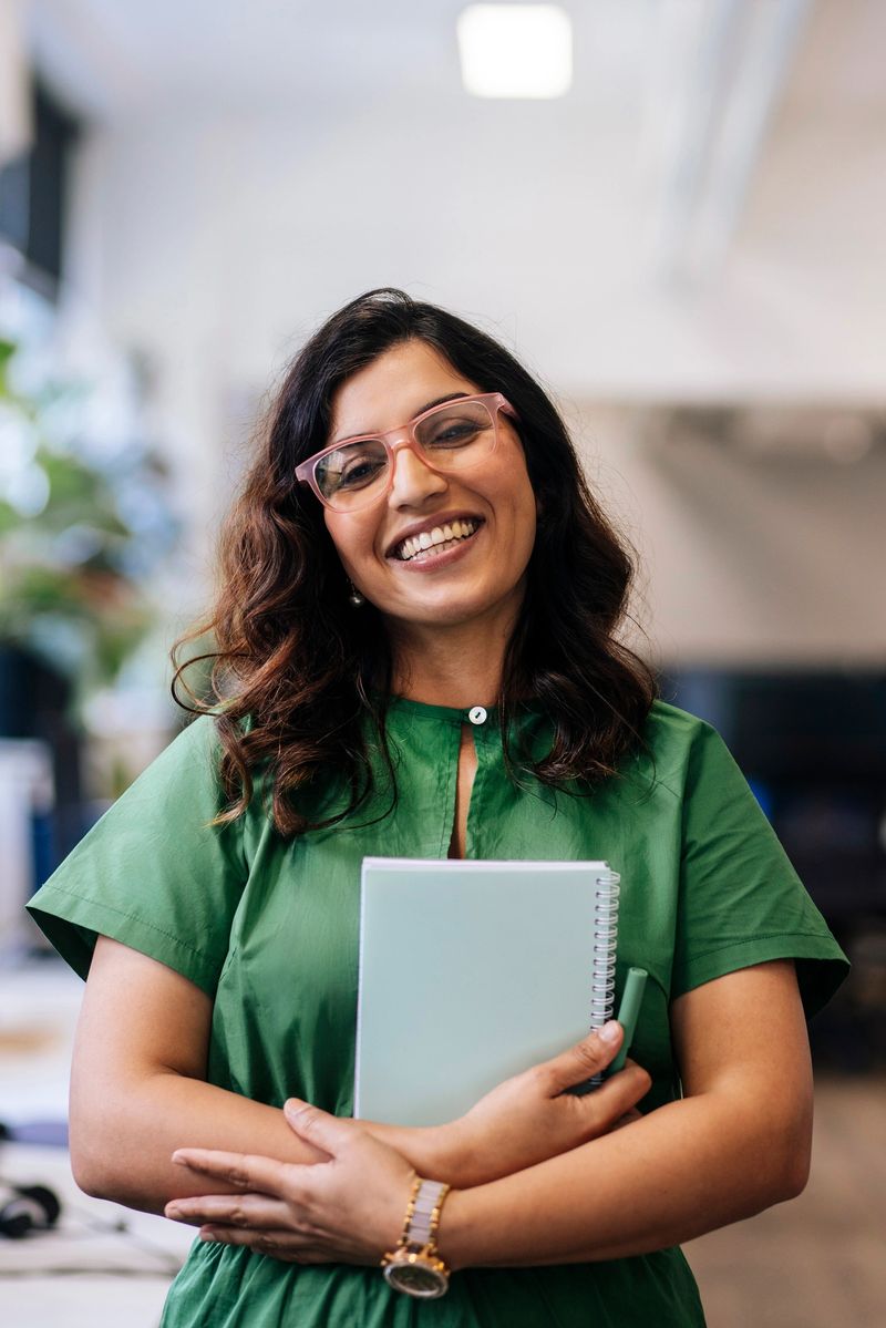 Portrait confident businesswoman with glasses and a warm smile, dressed in a green outfit, holds a notebook in a bright modern office environment
