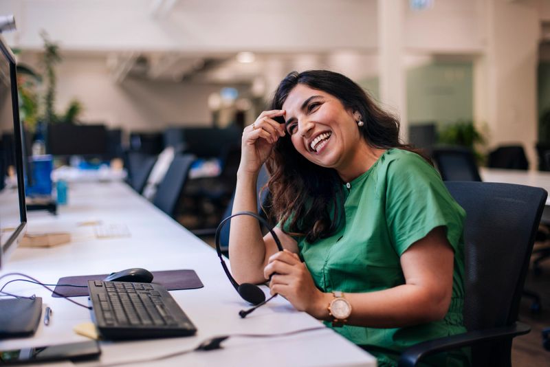 A cheerful customer support agent holds a headset in a modern call center office; She sits at a desk with a keyboard, smiling warmly
