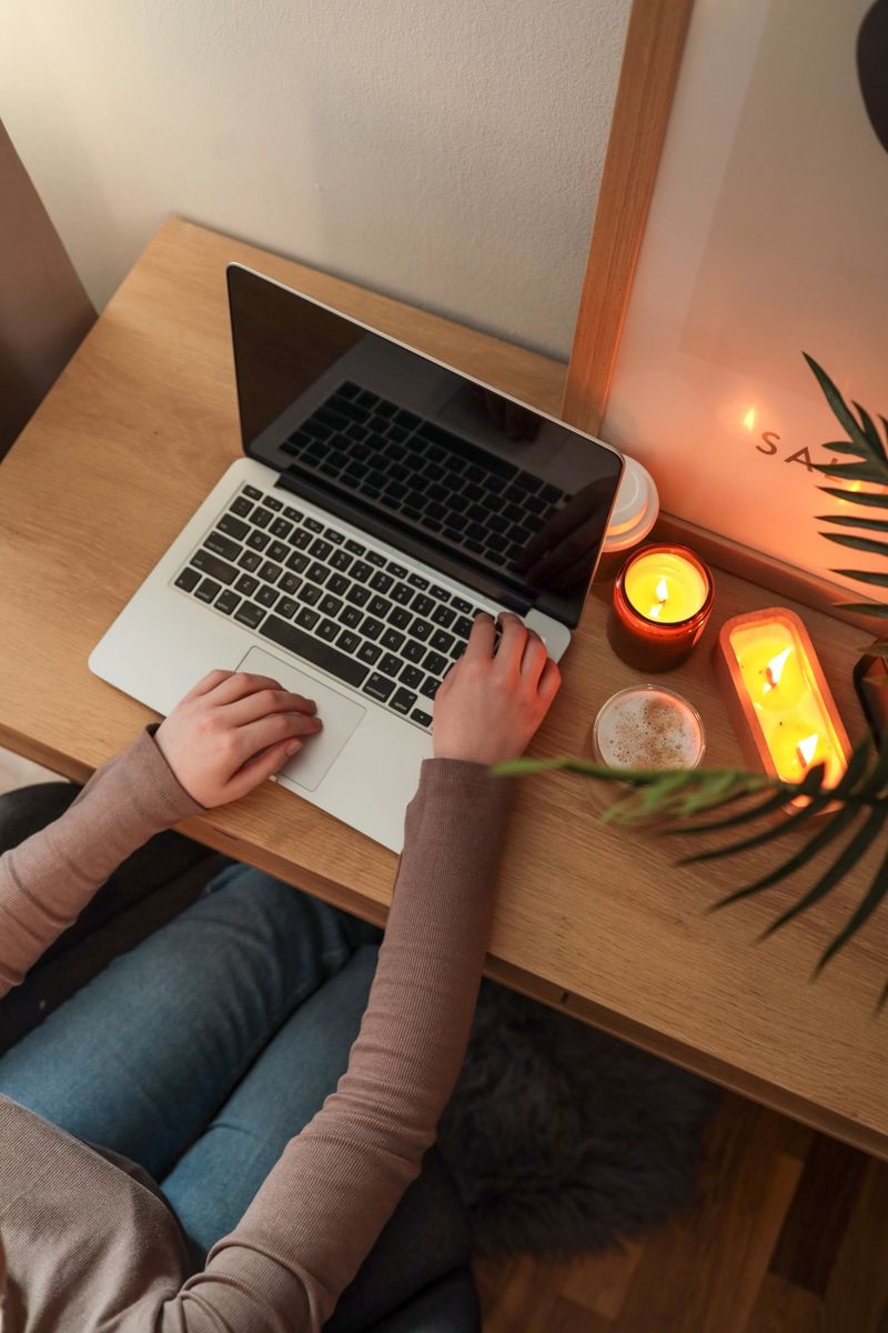 Top-down view of a person using a laptop at a wooden desk, with a cozy setup including lit candles, a cup of coffee, and decorative greenery. The warm ambient light creates a calm work atmosphere.