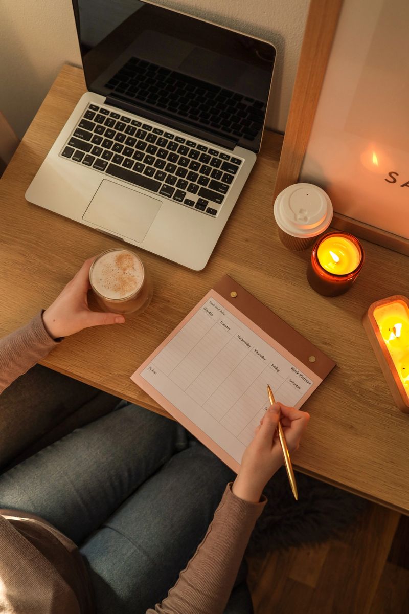 Top view of a person sitting at a wooden desk with an open laptop, holding a glass of coffee in one hand and writing in a weekly planner with a golden pen in the other hand.