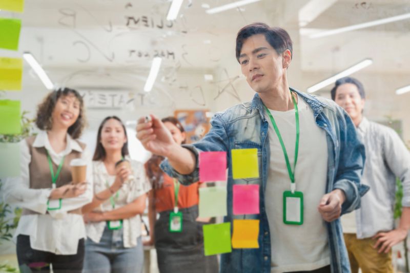 Asian business creative marketing team brainstorming startup strategy using glassboard and sticky notes. Collaborative ideation session demonstrates design thinking methodology disruptive innovation