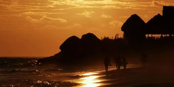 Silhouettes on beach at sunset with huts in background.