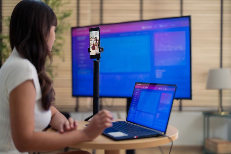 Asian woman leads online selling team using laptops and monitors, managing orders behind broadcast production. Team supports influencer livestream commerce through real-time analytics and omnichannel fulfillment operations