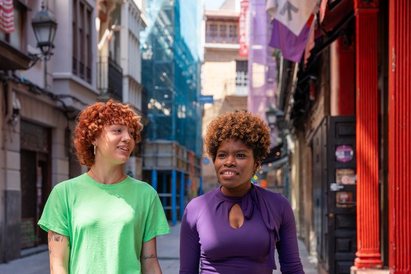 Two young women, one with red curly hair and the other with an afro, are enjoying a walk and conversation on a city street