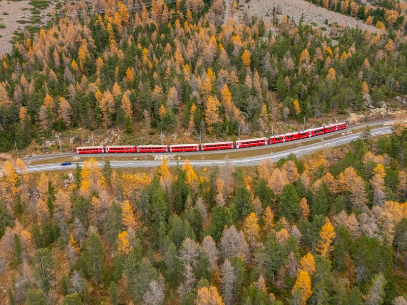 A red passenger train winds along tracks through a colorful autumn mountain forest. Scenic railway journey in fall with vibrant yellow and green trees. Mountain travel and nature landscape.