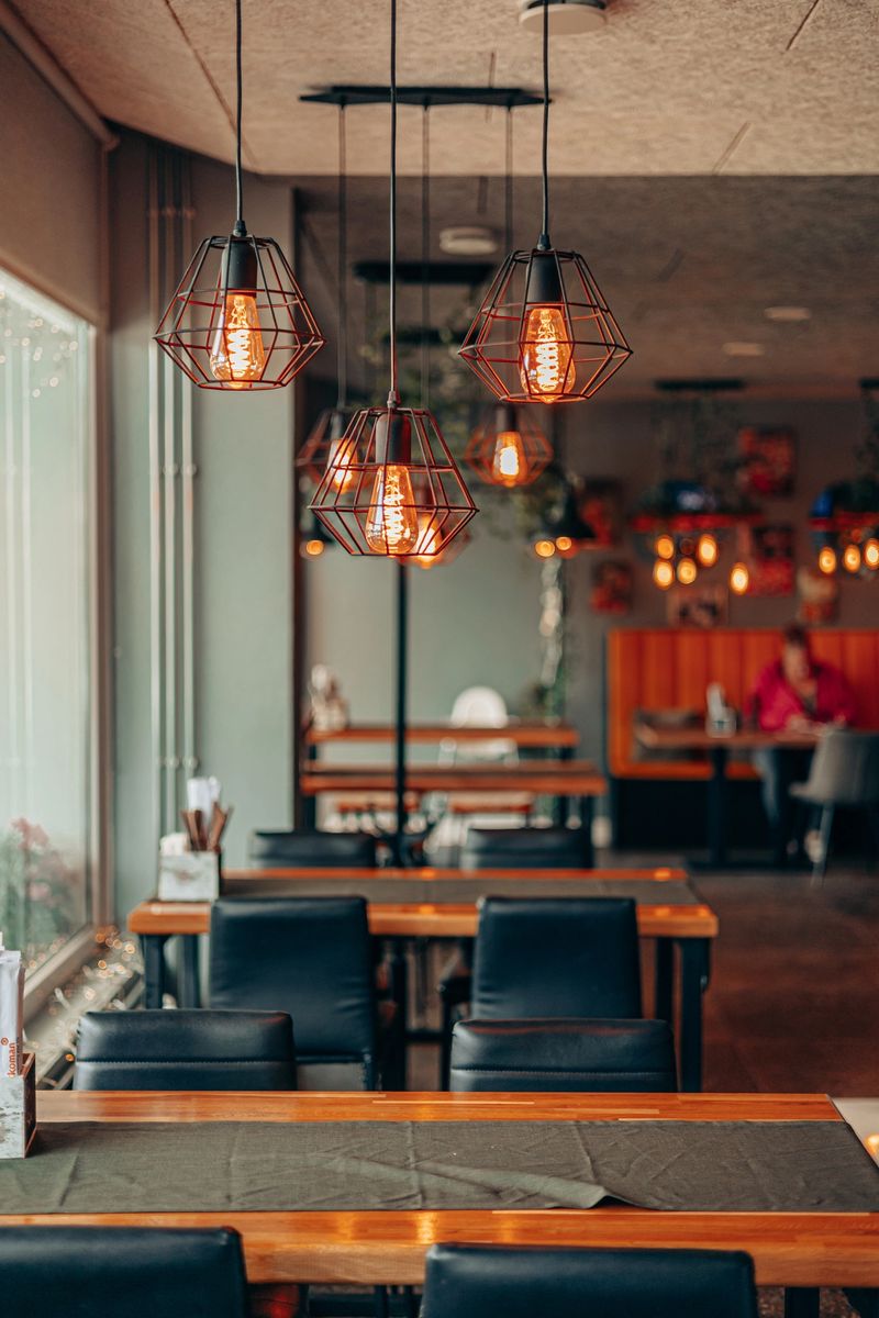 Warm industrial-style cage lamps with glowing Edison bulbs illuminate a modern cozy cafe interior, featuring wooden tables, black chairs, and a relaxed inviting atmosphere