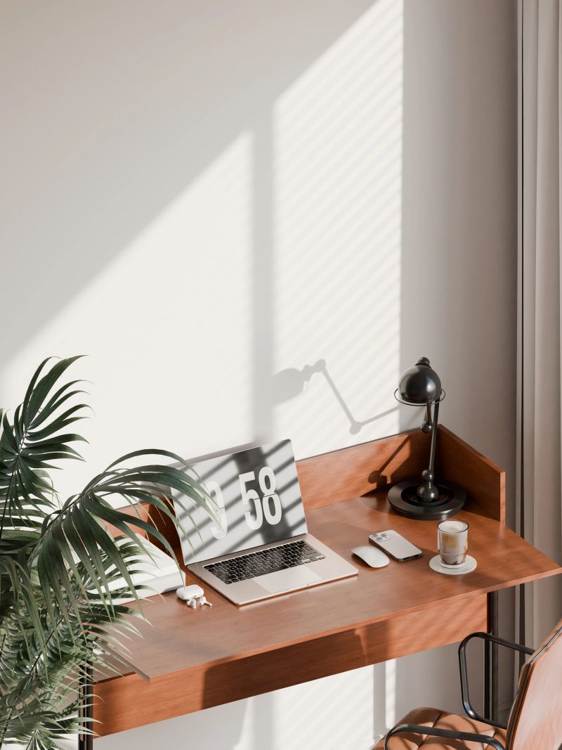 Modern home office interior with a minimalist design featuring a wooden desk, ergonomic chair, and a vertical blank frame mockup on the wall. Bright natural light, green plant decor, and an organized workspace setup make it ideal for showcasing poster designs, wall art, or home office inspiration.