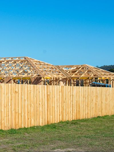 Wooden house frames under construction behind a tall wooden fence on a sunny day.