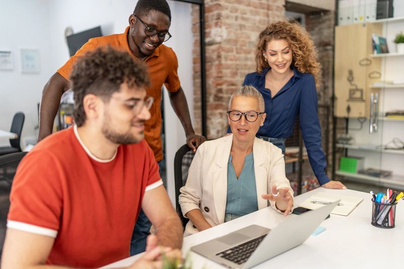 Diverse team of business professionals is gathered around a laptop, engaged in a productive discussion, sharing ideas, and working together in a modern office environment
