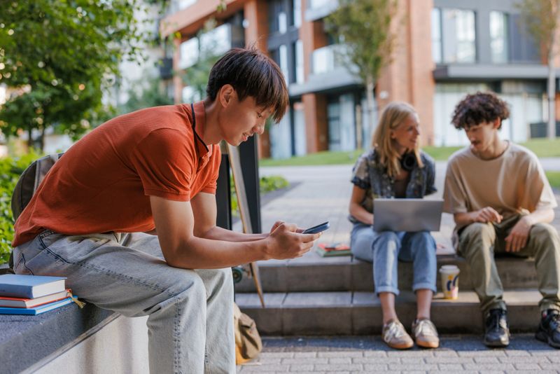 Young student using smart phone, while two other are talking and using a laptop outdoors.