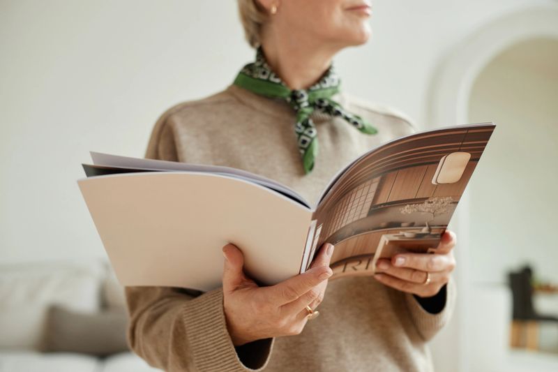 Caucasian senior woman holding open magazine with interior design images, standing indoors with partial face visible, wearing scarf around neck, reading content attentively