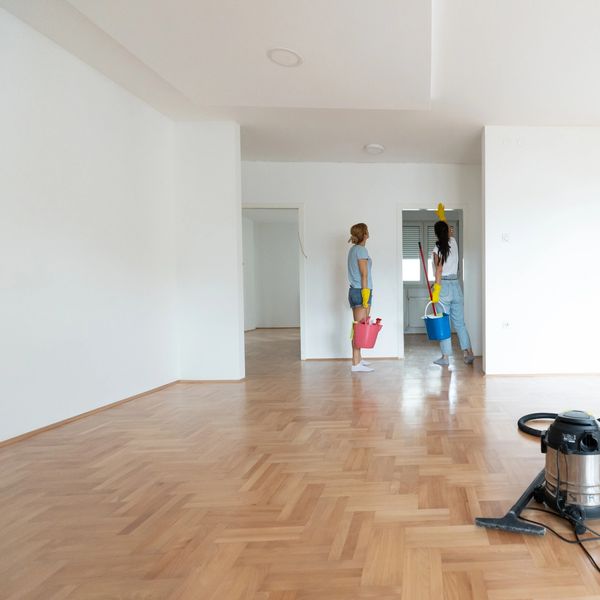 Two women cleaning a spacious empty room with wooden flooring.