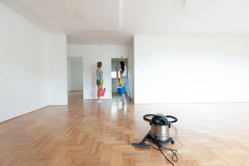 Two individuals are cleaning an empty, white-walled room with wooden floors using cleaning supplies, including buckets and a vacuum cleaner. A sense of organization and tidiness is emphasized.