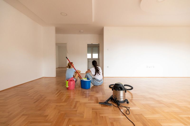 Two women seated in a freshly cleaned empty apartment, surrounded by cleaning tools and supplies, visible vacuum and bright interior.
