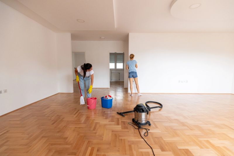 Two women engage in thorough cleaning of an empty minimalist room with wooden flooring. Various cleaning supplies, such as buckets and a vacuum, are seen, emphasizing cleanliness and preparation for new occupants.