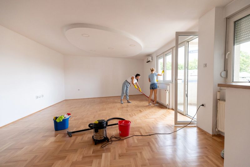 Two individuals are cleaning an empty apartment, focusing on windows and floors using cleaning supplies. The bright space emphasizes a fresh, organized atmosphere suitable for moving in or professional cleaning services.