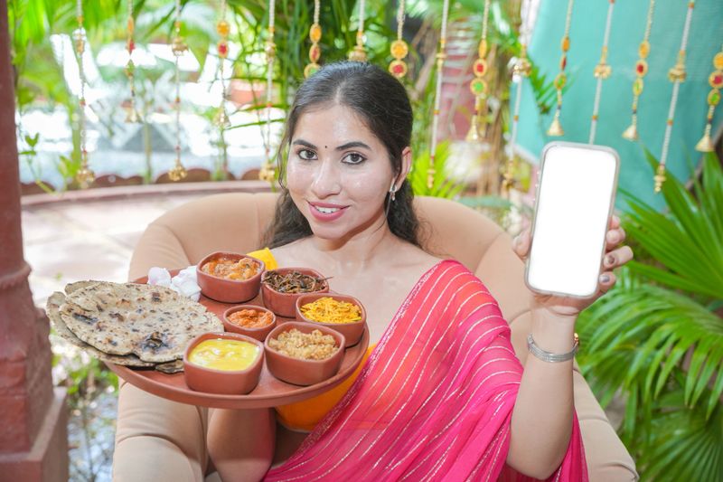 Young woman holding plate full of north indian vegetarian food platter or rajasthani thali show blank smart phone display scree for advertisement. Complete meal. Restaurant menu. India.