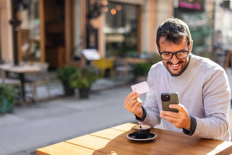 Mid adult man smiling while holding a credit card and using a smartphone for online shopping at a cafe, enjoying the convenience of digital payments in a vibrant urban setting
