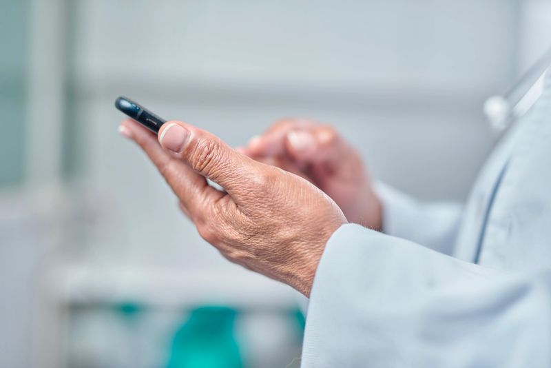 Close-up view of a medical worker's hands holding a smartphone, highlighting technology use in healthcare settings and communication. The background conveys a professional, clean clinic or office environment.