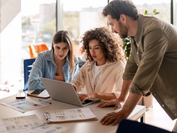 Three young professionals collaborate over a laptop in a bright office.