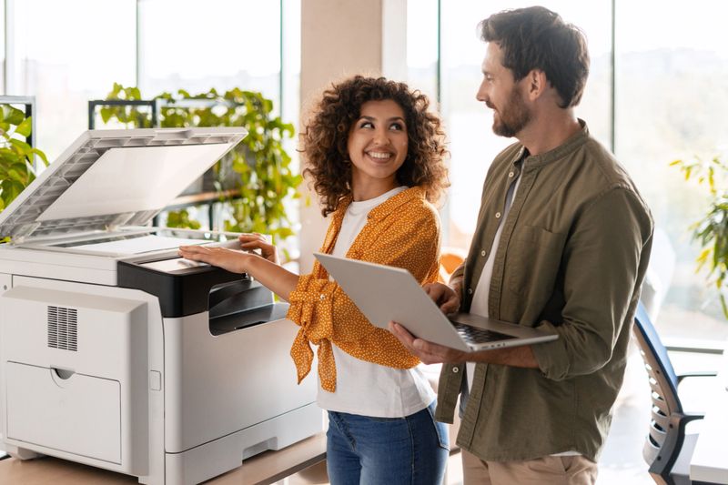 Two smiling colleagues collaborating while using a multifunction printer and a laptop in a bright, modern office environment
