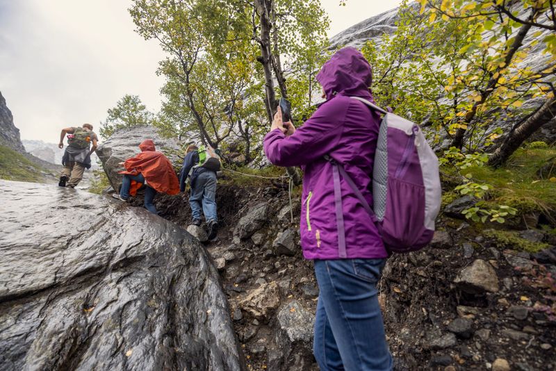 Hikers family navigates a steep, rain-slicked rocky section of the trail leading to the Buarbreen glacier, an outflow of the larger Folgefonna glacier in Norway. One of the hikers - a mature woman is taking photos of the teenage kids gripping a safety chain bolted into the rock for support. The terrain is rugged and damp, with patches of vegetation clinging to the rocks, reflecting the challenging conditions of hiking in wet weather. 
Shot with Canon R5 on an overcast summer day.