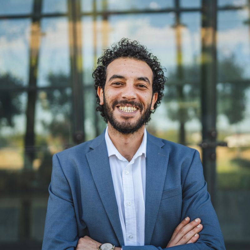 Confident businessman in suit standing with arms crossed, smiling outside of modern office building, projecting success and professionalism