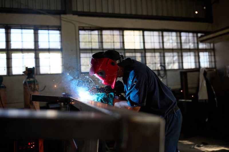 Welder wearing safety equipment works on steel fabrication inside a well-lit industrial workshop. The image showcases precision welding techniques in action.