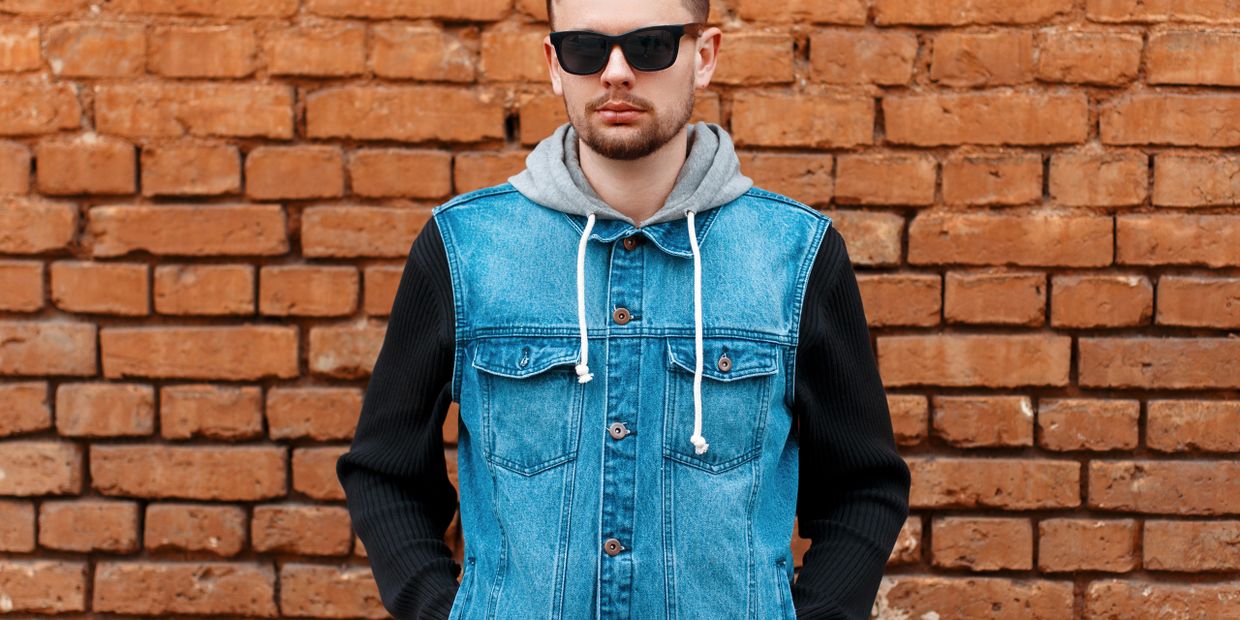 Man wearing sunglasses and a denim jacket with a gray hood stands against a brick wall.