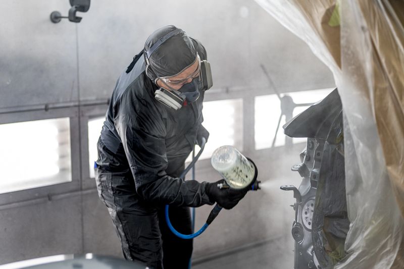 An automotive specialist in protective gear is applying a spray coat to a car panel in a workshop. The individual is using a spray gun, focusing intently on the task, featuring a controlled environment with surrounding plastic sheeting.