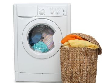 White front-load washing machine with colorful clothes inside and a wicker laundry basket overflowing with garments.