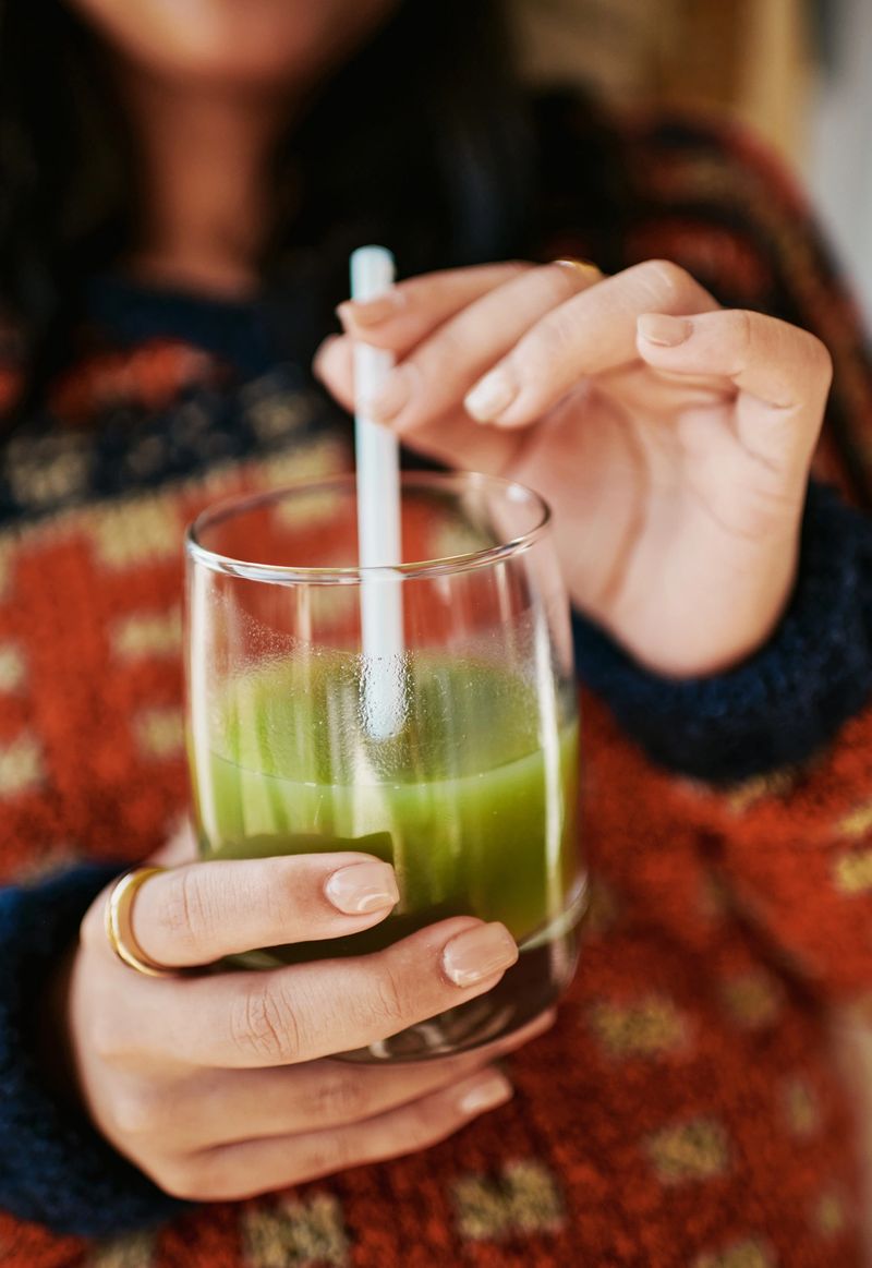 A close-up of a woman?s hands holding a glass of vibrant green juice. She wears a warm, patterned sweater, creating a cozy and inviting atmosphere.