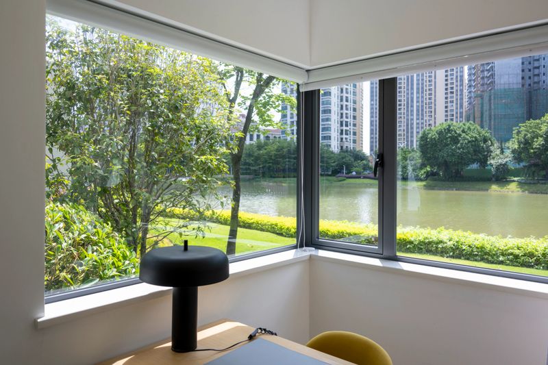 A serene office space with large windows overlooking a lush green landscape and a body of water. The minimalist design features a desk, chair, and decorative item, creating a peaceful work environment.