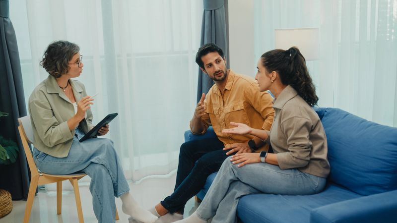 Young couple in serious discussion displeasure with mental health professional during marriage counseling session sit on sofa in living room at home. Therapy session and mental health concept.