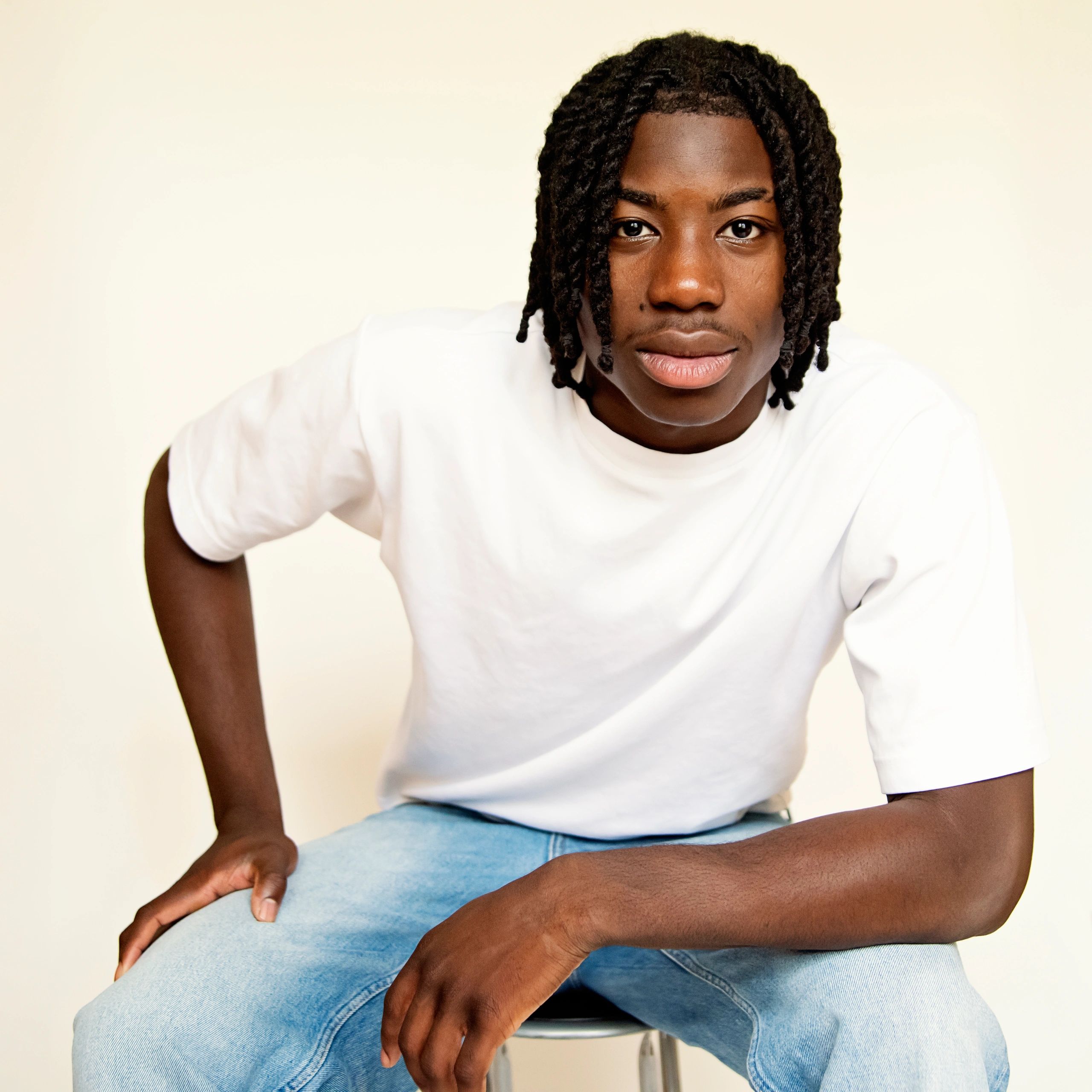 Young man with braided hair in white shirt and blue jeans posing.