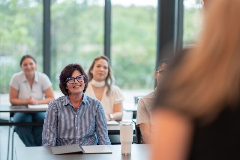 An engaged group of diverse adult learners attends an educational class, focused and smiling, in a bright classroom.