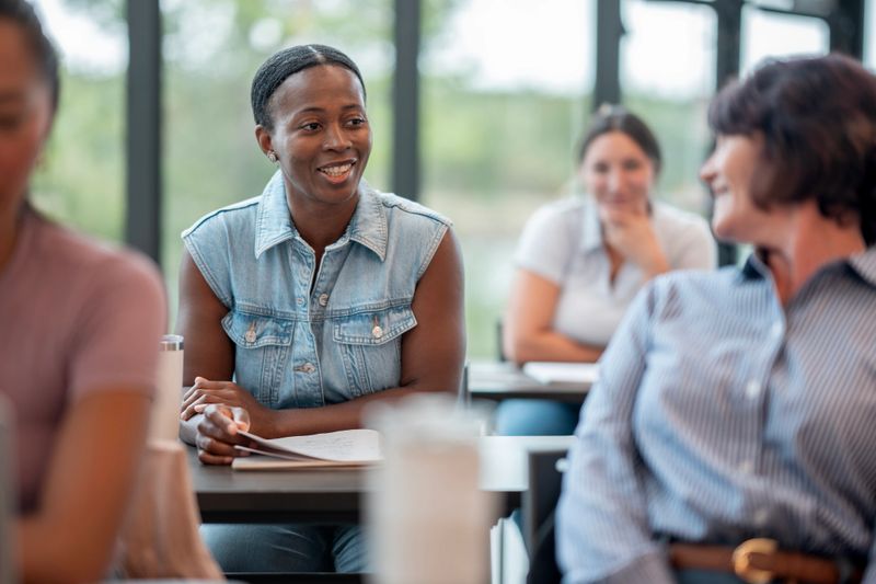 A diverse group engages in a collaborative discussion within a bright, modern classroom. Joyful and friendly interactions highlight teamwork. Participants appear attentive and involved in sharing ideas and learning from one another.