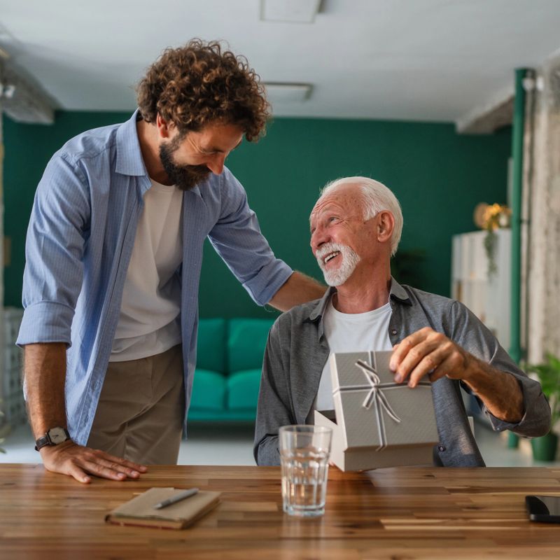 Adult son joyfully giving a thoughtful gift to his senior father at home, capturing the essence of love and care in their family relationship during a special celebration