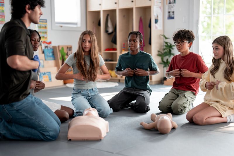 Group of kids participating in a CPR training activity led by an instructor inside a classroom environment, demonstrating life-saving techniques using training dummies, emphasizing skill development and teamwork.