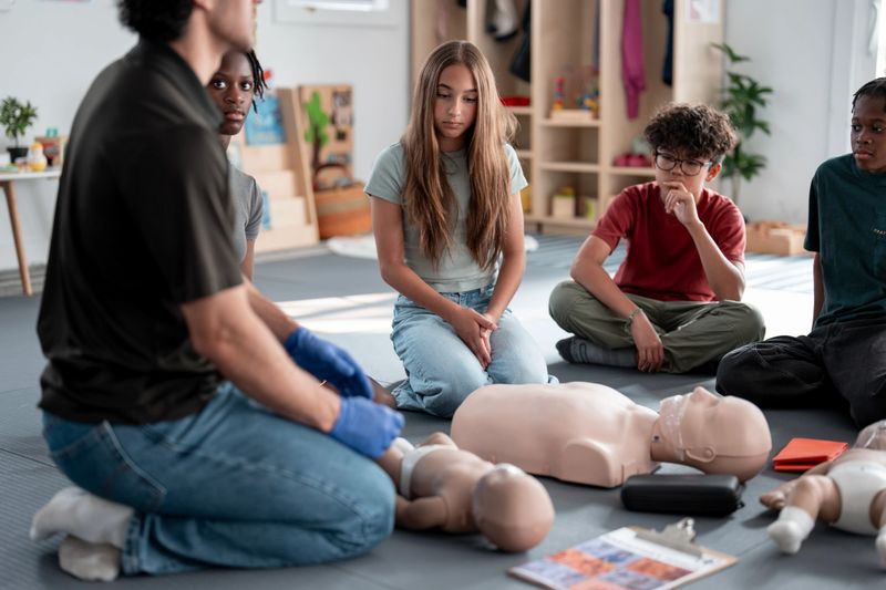 A group of diverse teenagers attentively learning CPR techniques from an instructor using a dummy in a classroom setting, focusing on essential lifesaving skills and first-aid training.