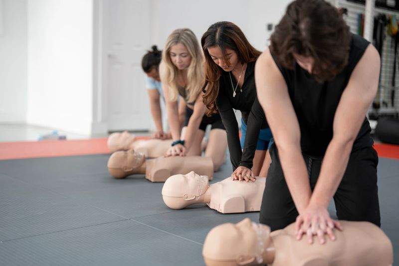 A group of diverse individuals are practicing cardiopulmonary resuscitation (CPR) on mannequins in an indoor classroom setup. The session emphasizes teamwork and lifesaving skills training in a professional environment.